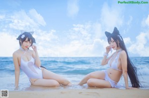 Two women in white bathing suits sitting on a rock by the ocean.
